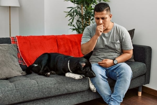A man coughing into his fist while sitting on his couch besides his dog.