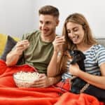 A couple on the couch with a red blanket, a bowl of popcorn, and a hungry Dachshund.