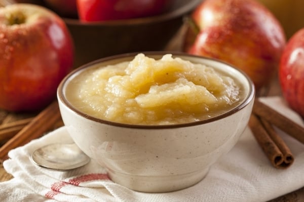 Fresh applesauce in a bowl surrounded by cinnamon sticks and whole apples.